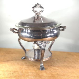 Può includere: Una chafing dish argentata con inserto e coperchio in vetro. Il piatto ha manici decorati e una maniglia decorativa sul coperchio. Poggia su un supporto argentato con gambe curve e una base riscaldante, ideale per buffet.