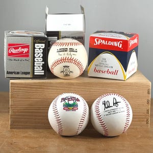 May include: A collection of baseballs and boxes. The boxes are labeled "Rawlings American League Baseball" and "Spalding Little League Baseball." Two baseballs are on the bottom, one with a team logo and the other signed.