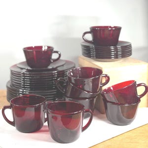 Puede incluir: Un conjunto de vajilla de vidrio rojo oscuro que incluye varios platos y platillos apilados, varias tazas a juego con asas y una jarra pequeña. La cristalería presenta un tono rubí oscuro translúcido y un diseño redondeado.