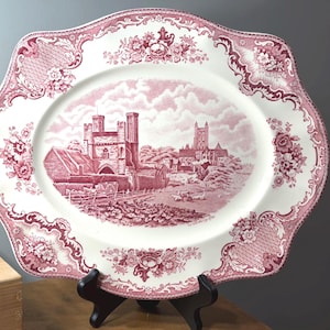 May include: An oval, antique serving platter with a red and white design. The center features a detailed landscape scene of a building with towers. The border has a floral and scroll pattern.