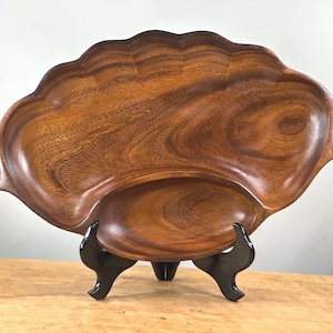 Puede incluir: Una gran bandeja de madera para servir con un borde festoneado y dos compartimentos integrados. La bandeja está soportada por un soporte negro. La veta de la madera es visible, mostrando un color marrón cálido. La bandeja está diseñada para servir alimentos.