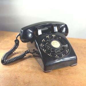 Może przedstawiać: Zabytkowy czarny telefon z tarczą obrotową z słuchawką i zwiniętym kablem. Tarcza posiada cyfry i litery, widoczny jest numer telefonu 649-0505. Telefon stoi na drewnianej powierzchni.