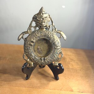 May include: A circular brass ashtray featuring an intricate relief of a mythical figure with a fierce expression at the top. The outer rim is decorated with detailed patterns and small figures, while the center is a smooth, recessed basin.