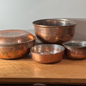 May include: Four copper bowls of varying sizes sit on a wooden surface. The largest bowl has a lid with an embossed design. The other bowls are open, revealing their interiors. The copper has a warm, reddish-brown hue.