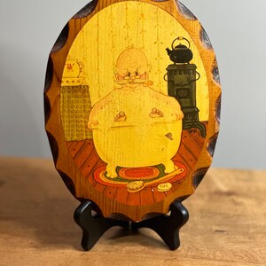 Peut inclure: Une plaque en bois avec une image peinte d'un homme dans un bain. L'homme a une barbe et une moustache et porte une serviette. La plaque est de forme ovale et a un bord festonné. La plaque est sur un support noir.