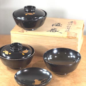 May include: A collection of black lacquerware bowls and lids with gold detailing. The set includes a covered bowl, a bowl with a lid, a small bowl, and a small plate. The bowls are displayed on a wooden surface.