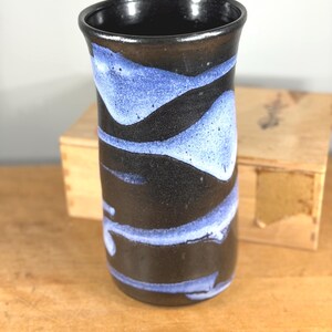 May include: A black ceramic vase with blue wave patterns. The vase has a cylindrical shape and a glossy finish. The interior is black. The vase is on a wooden surface. The vase is likely handmade.