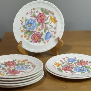 Puede incluir: Un juego de cinco platos de cerámica blanca con un diseño floral con flores rosas, azules y amarillas. Los platos están apilados uno encima del otro.