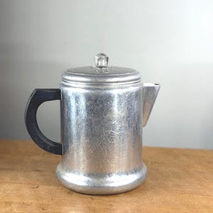 Può includere: Una caffettiera vintage in alluminio con un manico nero e un pomello trasparente sfaccettato sul coperchio. La caffettiera ha un beccuccio e una base arrotondata. Il metallo ha una finitura spazzolata e la caffettiera è su una superficie di legno.