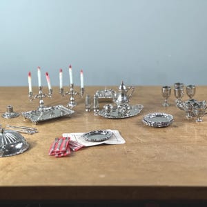 Puede incluir: Juego de comedor plateado en miniatura con una variedad de piezas, que incluyen un candelabro con velas de punta roja, bandejas, una tetera y copas. El juego también incluye una fuente cubierta, platos y una servilleta.