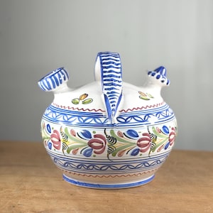 May include: A white ceramic pitcher with a handle and two spouts. The pitcher is decorated with blue, red, and green floral patterns and blue stripes. The pitcher is sitting on a wooden surface.