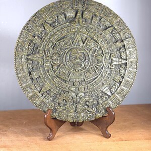 Könnte beinhalten: Eine detaillierte, kreisförmige Nachbildung des Maya-Kalenders in Gold- und Grüntönen, präsentiert auf einem Holzständer. Das komplizierte Design zeigt verschiedene Symbole und Figuren, die alte Kunstfertigkeit zeigen.