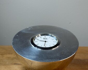 May include: A silver clock with a white face and black hands. The clock is on a wooden surface. The clock face says "Studio".