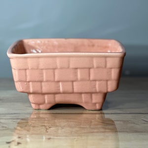 Puede incluir: Un macetero de cerámica de color rosa pálido con forma cuadrada y un patrón de ladrillo tejido. El macetero tiene cuatro pequeñas patas.