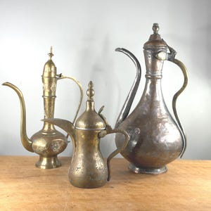 Może przedstawiać: Trzy antyczne metalowe dzbany o różnych rozmiarach i wzorach. Dzbany po lewej stronie są w kolorze złotym z wydłużonym dziobkiem. Środkowy dzban jest mniejszy, z uchwytem i pokrywką. Największy dzban jest w kolorze miedzi.