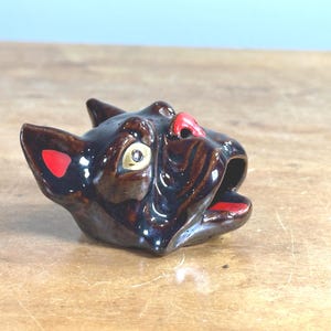 以下が含まれることがあります： 光沢のある濃い茶色の仕上げのセラミック製の猫の頭の置物。猫は耳と口に赤いアクセント、赤い鼻、金色の目があります。置物は装飾品です。