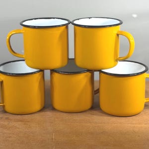 May include: A set of five bright yellow enamel mugs with white interiors and black rims. Each mug features a smooth, glossy finish, a sturdy side handle, and a cylindrical body. The mugs are arranged in a stacked formation, showcasing their uniform design and vibrant color.