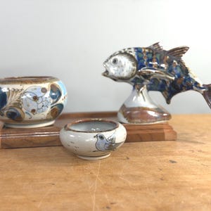 Puede incluir: Colección de objetos decorativos de cerámica, que incluye una escultura de pez y dos cuencos pequeños. El pez es azul, marrón y blanco, y los cuencos presentan diseños florales y de pájaros. Los objetos se exhiben en una bandeja de madera.