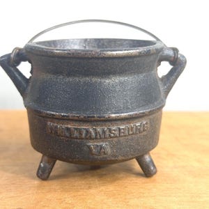 Könnte beinhalten: Ein kleiner, dunkelgrauer Gusseisenkessel mit drei kurzen Beinen, zwei seitlichen Griffen und einem dünnen Drahtbügelgriff. Auf der Vorderseite des Topfes befindet sich der geprägte Text "WILLIAMSBURG" in der oberen und "VA" in der unteren Zeile.
