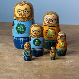 May include: Set of six nesting dolls, each painted with a bearded man wearing glasses and a blue shirt with the text "1% for Peace". The dolls are painted in shades of blue, orange, and green.