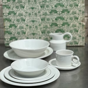 May include: A set of white ceramic dishes, including a large bowl, a smaller bowl, a plate, a saucer, and a mug. The dishes are arranged on a silver surface.