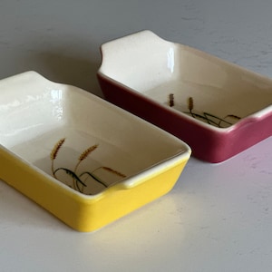 May include: Two small ceramic baking dishes with handles. The dish on the left is yellow with a wheat design. The dish on the right is pink with a wheat design.