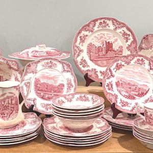 Puede incluir: Un conjunto de vajilla antigua de porcelana rosa y blanca, que incluye platos, cuencos, tazas y piezas para servir. Las piezas presentan un diseño paisajístico con un castillo y un borde floral. El conjunto incluye varios tamaños y formas, ideal para una cena formal.