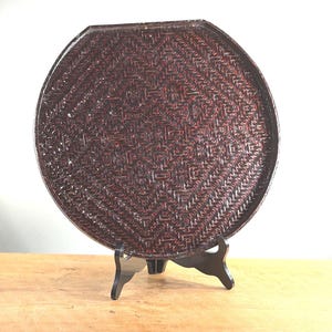 May include: A round, dark brown woven tray with a geometric pattern, displayed on a black stand. The tray's surface has a textured appearance, and the stand has a three-legged design. The tray is likely made of natural materials.