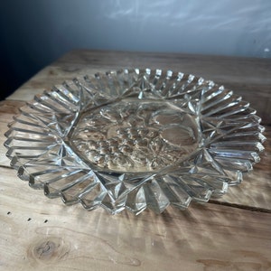Puede incluir: Un plato de servir de vidrio transparente con un diseño en relieve de uvas y hojas. El plato tiene un borde festoneado y un patrón de diamante.