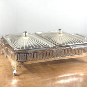 Könnte beinhalten: Ein silberfarbenes Chafing-Dish mit zwei Fächern, Glasdeckeln und Griffen. Das Gericht hat ein dekoratives, geriffeltes Design auf den Deckeln und einen detaillierten, verzierten Sockel mit kleinen Füßen. Zum Servieren von Speisen.