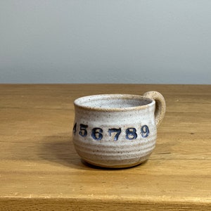 Könnte beinhalten: Eine kleine, weiße Keramiktasse mit Henkel. Die Tasse hat eine gesprenkelte Glasur und die Zahlen 456789 sind auf der Seite aufgedruckt.