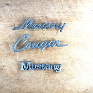 以下が含まれることがあります： シルバーグレーの金属仕上げの「Mercury」、「Cougar」、「Mustang」の3つのヴィンテージカーエンブレム。 エンブレムは、クラシックなデザインと自動車の伝統を示す、明るい茶色の木製の表面に表示されています。