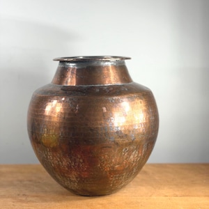 May include: A round, hammered copper vessel with a wide, bulbous body and a narrow, flared rim. The surface displays a rich, metallic brown patina with visible texture from the hand-hammering process, creating a reflective and aged appearance.