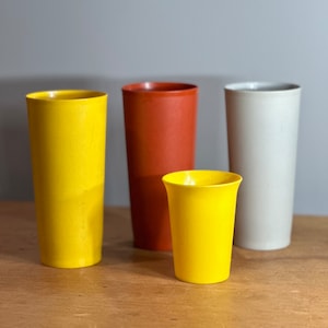 May include: Four plastic cups in different colours: yellow, orange, white, and a smaller yellow cup. The cups are on a wooden surface.