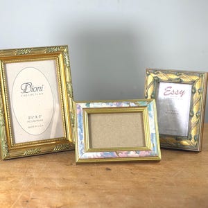 May include: Three decorative picture frames in gold tones. The frames vary in size and design, with ornate detailing and floral patterns. One frame has the text "Dioni Collection" and another has "Essy". The frames are designed to hold 3" x 5" pictures.