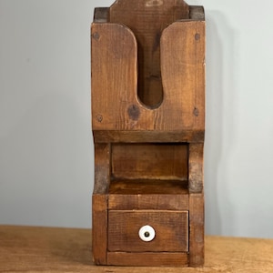 Puede incluir: Una unidad de almacenamiento de madera rústica montada en la pared con un estante y un pequeño cajón. El cajón tiene una perilla blanca. La unidad está hecha de madera sin terminar y tiene un acabado de madera natural.