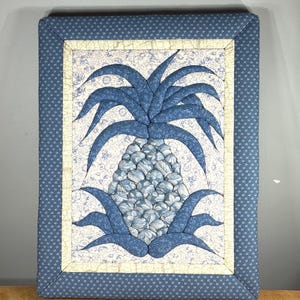 Puede incluir: Un tapiz rectangular de tela con un motivo central de piña construido con piezas de tela estampadas en azul y blanco. La piña se sitúa sobre un fondo floral claro con un borde crema y un marco exterior de color azul oscuro.