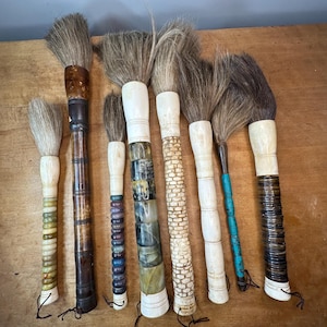 May include: A set of eight calligraphy brushes with various handle designs. The handles are made of wood, bone, and other materials. The brushes have natural bristles and are arranged on a wooden surface.