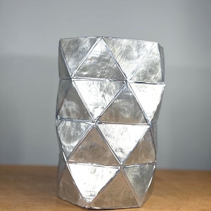 May include: A cylindrical vase constructed from multiple silver-colored triangular panels. The surface of each triangle features a brushed metallic texture, and the panels are joined together by thin, raised silver-colored seams, creating a geometric pattern.