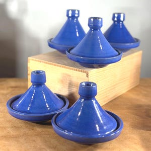 May include: A collection of cobalt blue ceramic tagine pots. Each pot features a conical lid and a circular base. The tagines are displayed on a wooden surface and a wooden box. The pots have a glossy finish and are designed for cooking and serving.