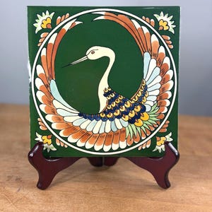 May include: A square ceramic tile featuring a dark green background with a central circular design of a white crane with outstretched wings. The crane's feathers are detailed in shades of orange, blue, yellow, and white, with floral accents in the corners.
