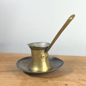 May include: A small, brass coffee pot with a long handle, resting on a silver-colored saucer. The pot has a rounded body and a flared top. The handle is attached with a small rivet. The saucer has a beaded edge. The items are on a wooden surface.