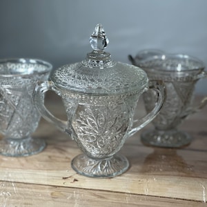 May include: A vintage glass sugar bowl and creamer set with a decorative, embossed pattern. The set features a lidded sugar bowl with two handles and a creamer with a single handle.
