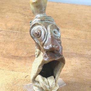 May include: A handcrafted ceramic sculpture with a stylized face, featuring large eyes, a prominent nose, and an open mouth with a protruding tongue. The sculpture has a textured surface with brown, tan, and green hues, and a cylindrical top.