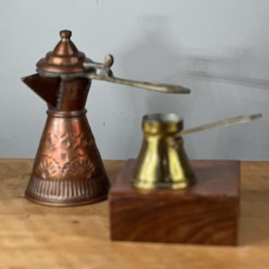 May include: A set of two vintage copper and brass coffee pots. The larger pot is copper with intricate floral designs and a handle. The smaller pot is brass with a simple design. Both pots are on a wooden base.