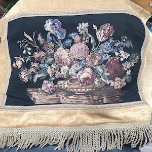 Puede incluir: Un tapiz con un bodegón floral en un jarrón, con iris, tulipanes y otras flores en tonos rosas, morados y azules. El tapiz tiene un fondo negro, un borde dorado y una borla dorada.