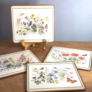 Op de afbeelding: Set van vier rechthoekige placemats met bloemen- en vlindermotieven. Elke placemat heeft een crèmekleurige achtergrond met een rand en een gedetailleerde illustratie van wilde bloemen en vlinders. Eén placemat wordt op een houten ezel tentoongesteld.
