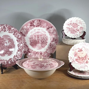 May include: A collection of antique pink and white china dishes. The set includes large plates with floral and landscape designs, a covered serving bowl, and smaller plates. The dishes are displayed on wooden stands.