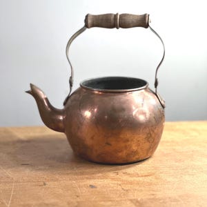 May include: A vintage copper kettle with a wooden handle and a curved metal handle. The kettle has a spout and a rounded body. The copper has a warm, reddish-brown colour, and the wood handle is light brown. The kettle is likely used for boiling water.