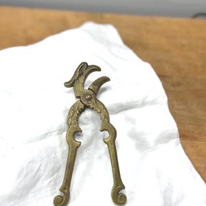 May include: A vintage brass nutcracker with a decorative bird-like design at the top. The handles have a curved shape with decorative swirls at the ends. The nutcracker is resting on a white cloth.